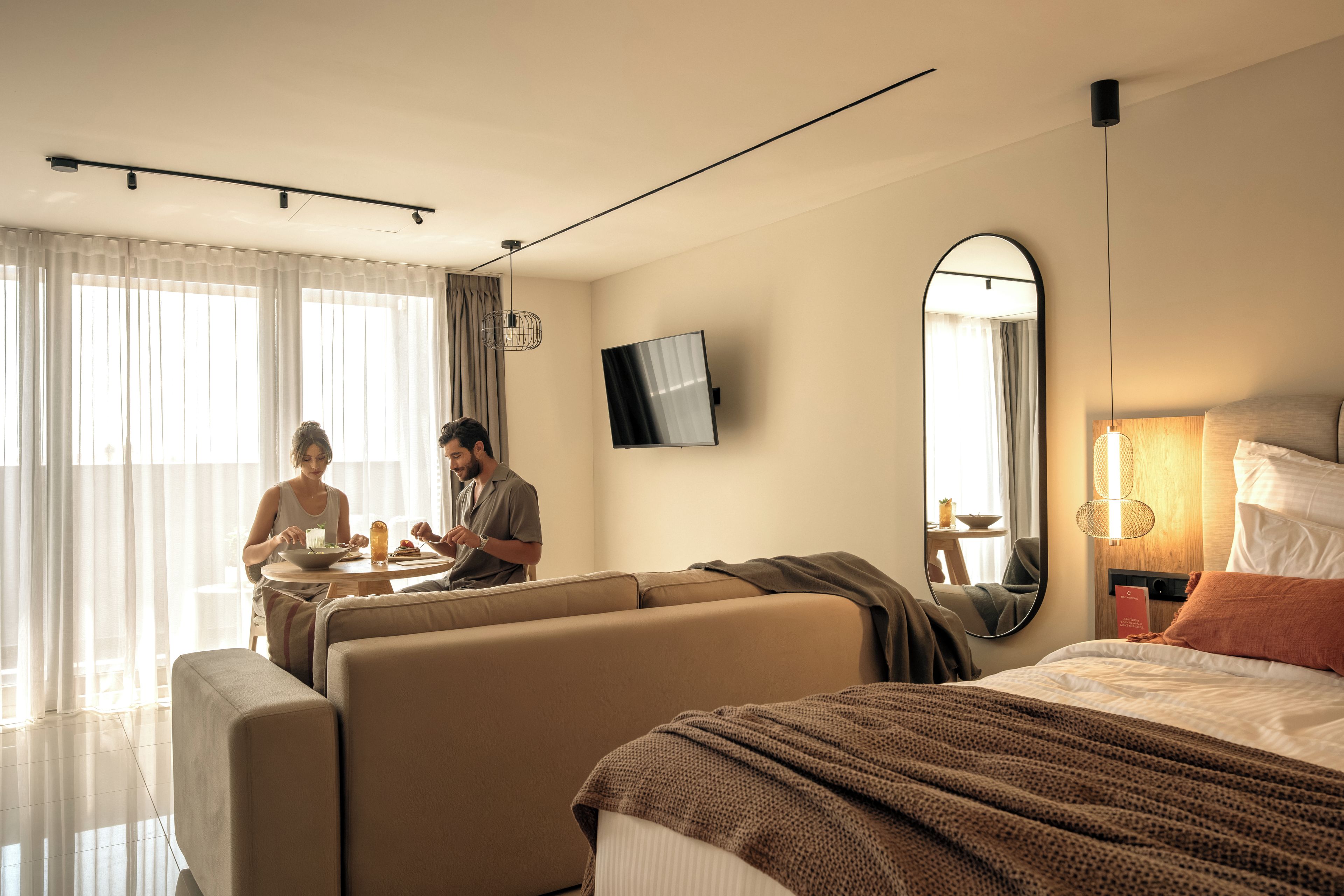 A hotel room with a bed, a sofa, a dining table, and a TV mounted on the wall.