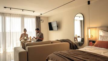 A hotel room with a bed, a sofa, a dining table, and a TV mounted on the wall.