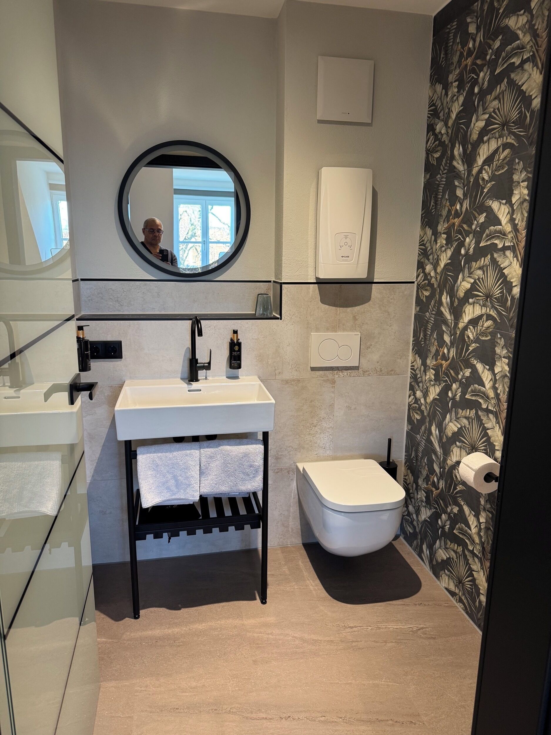 Un baño moderno con separador de vidrio, espejo redondo, lavamanos blanco, inodoro y zona de ducha.