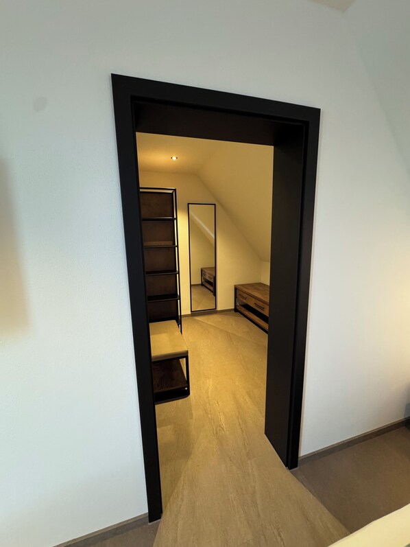 Un couloir étroit mène à une chambre dotée d’un plancher en bois, d’une armoire intégrée et d’un miroir au mur.
