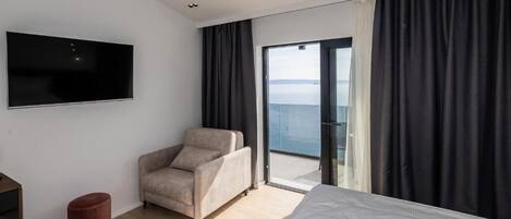 A modern hotel room with a large bed, a flat-screen TV mounted on the wall, a comfortable armchair, and a view of the sea through a sliding glass door.