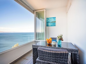 Ein Balkon mit Blick auf das Meer, ein Korbstuhl, ein Tisch, eine Vase mit Blumen und ein Gemälde.