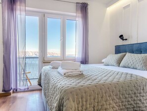 Ein Schlafzimmer mit Bett, Vorhängen am Fenster, einem Kopfteil und Blick ins Freie.