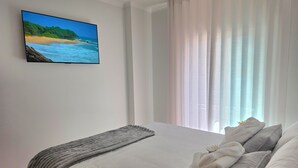 Un dormitorio con cama, televisor en la pared y ventana con cortinas traslúcidas.
