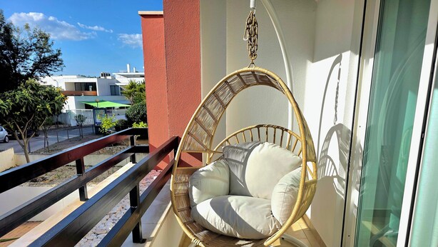 Una silla colgante de mimbre con cojines en un balcón con barandilla, puerta de cristal y vistas a edificios y árboles.
