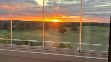 Quarto com uma grande janela com vista para o pôr do sol, uma cadeira de vime com almofada e paisagem verde ao fundo.