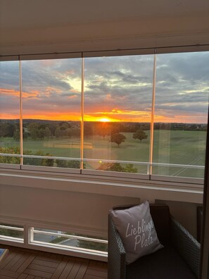 Quarto com uma grande janela com vista para o pôr do sol, uma cadeira de vime com almofada e paisagem verde ao fundo.