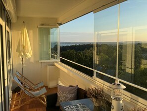 A balcony with a view of a forest and a lake, wooden furniture, a table with a lamp, and a cushion with text. - Ferienwohnung Lieblingsplatz, Urlaub und Meer! (Sierksdorf)