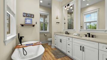 A modern bathroom with a white sink, vanity, and a large mirror. There are wooden floors, a window with blinds, and a shelf with decorative items.