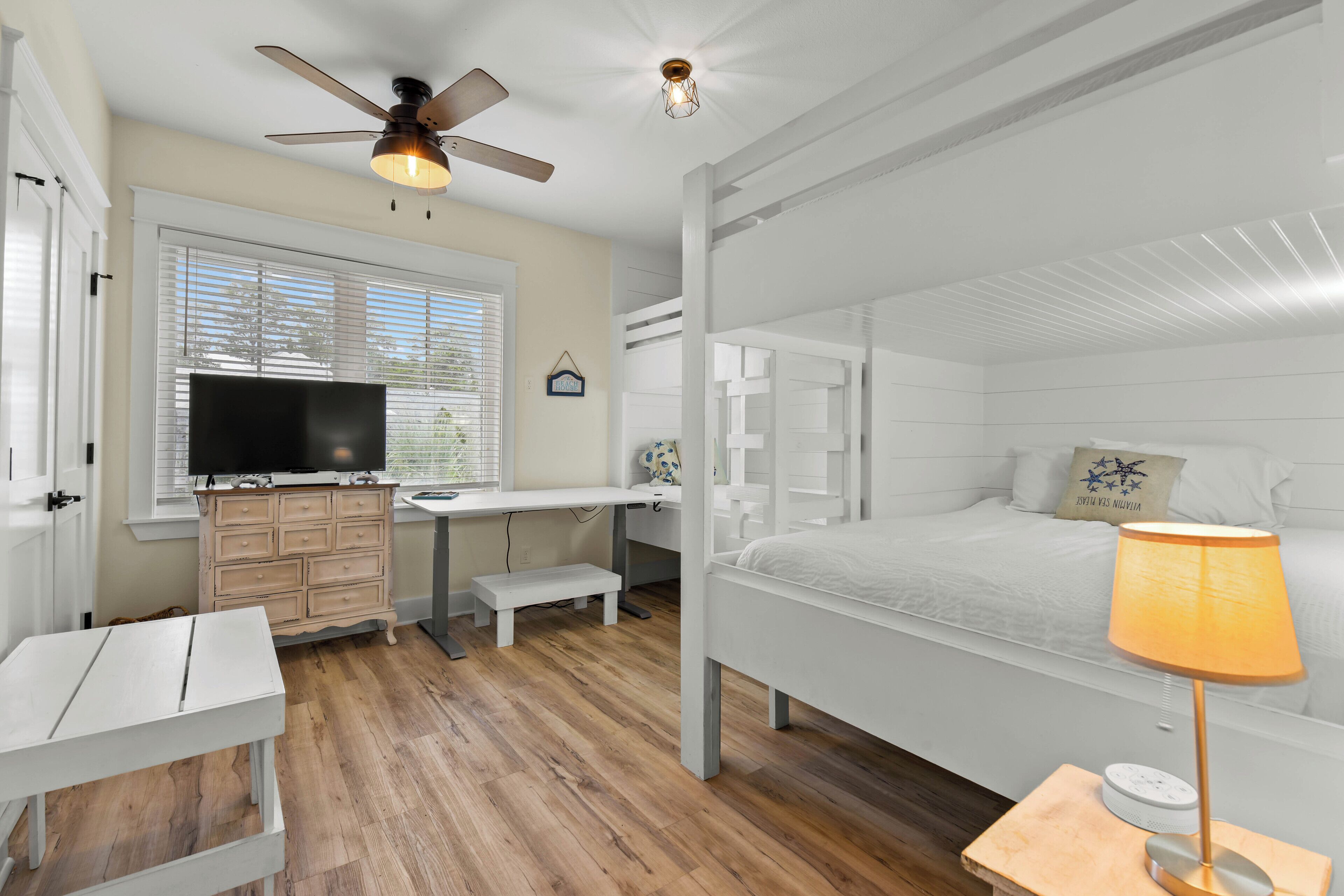 A bedroom with a bunk bed, a desk with a TV, a ceiling fan, and a bedside lamp.