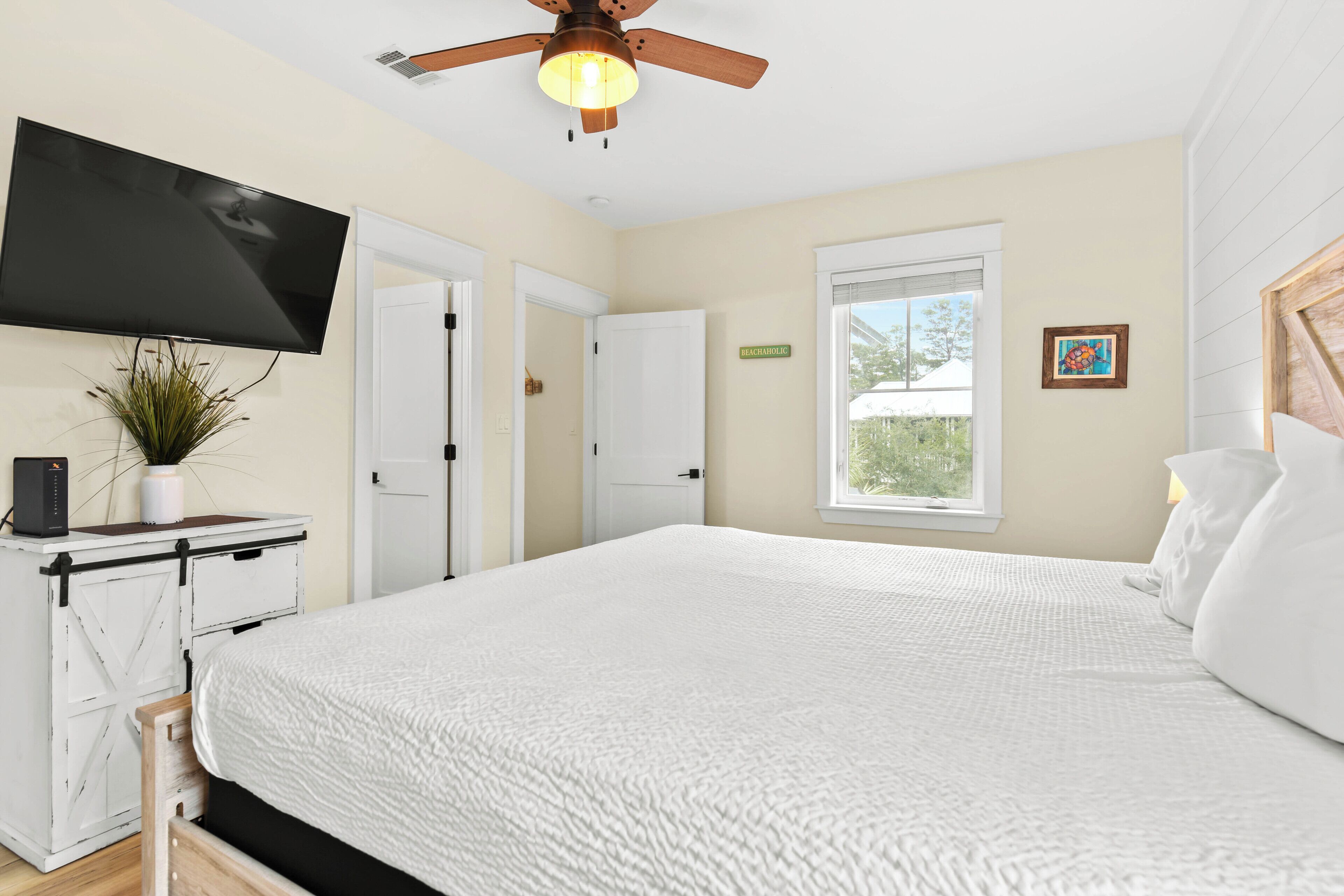 A bedroom with a bed, a ceiling fan, a TV mounted on the wall, and a window.