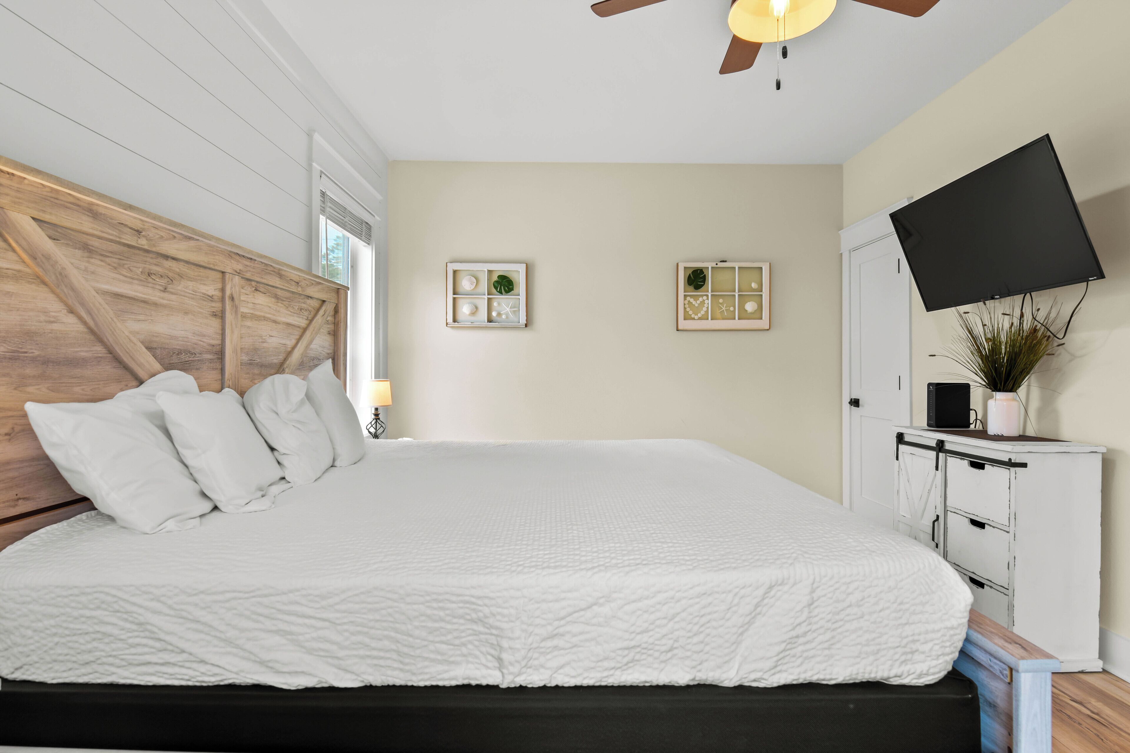 A large bed with a wooden headboard, a flat-screen TV mounted on the wall, and a small cabinet with a plant.