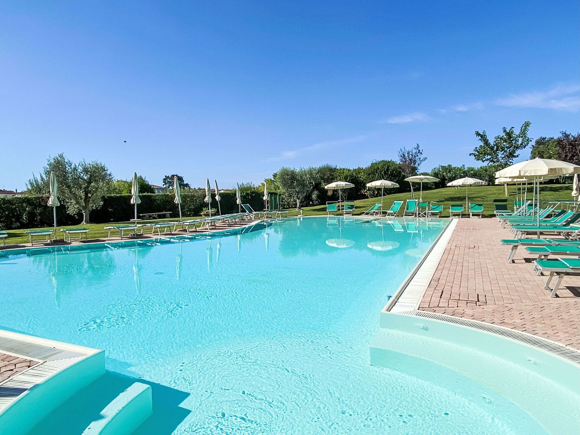 A clear blue swimming pool with sun loungers and umbrellas, surrounded by a brick deck and greenery.