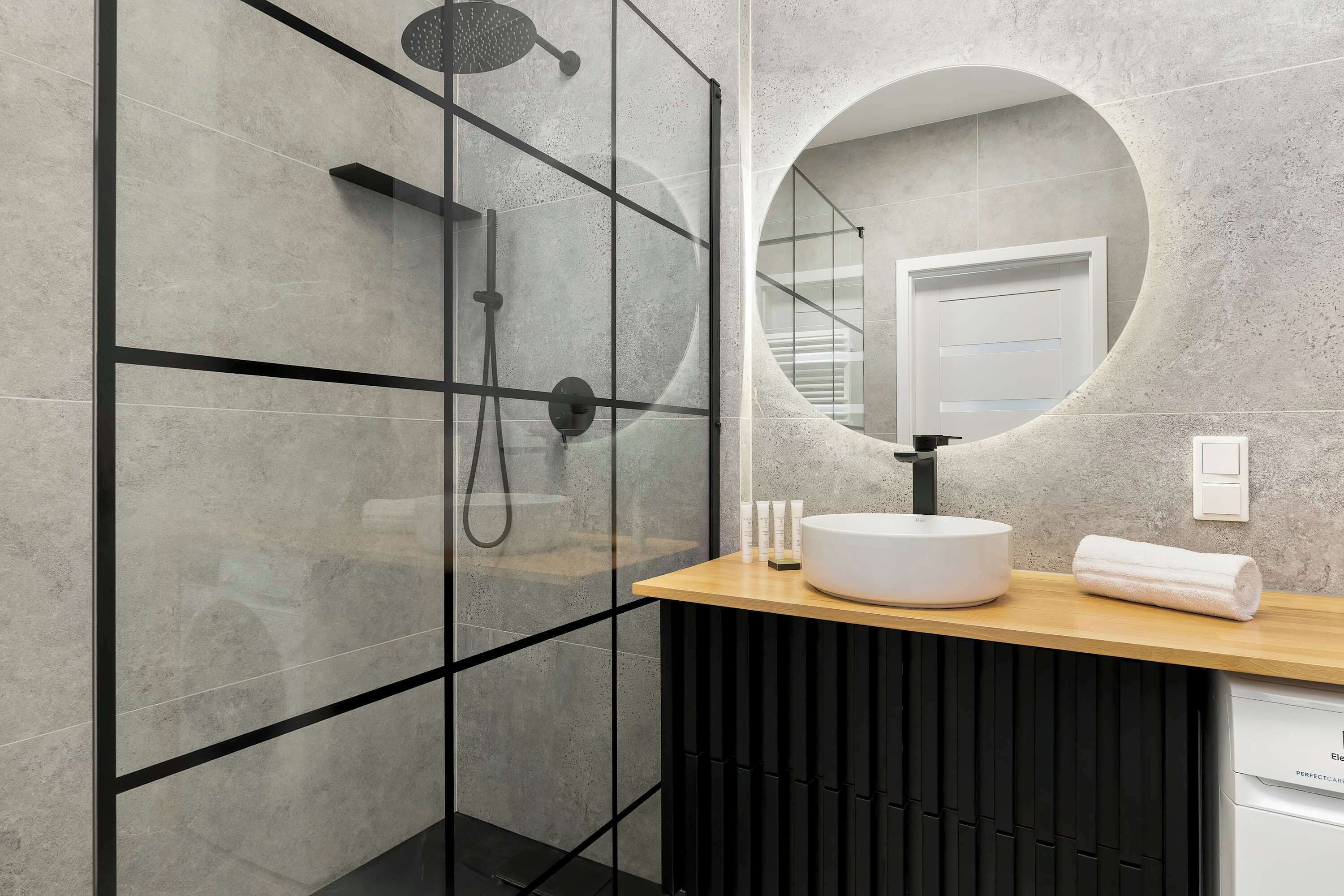 Een moderne badkamer met een douche in glazen cabine, een ronde spiegel en een houten wastafelmeubel met een witte wasbak.