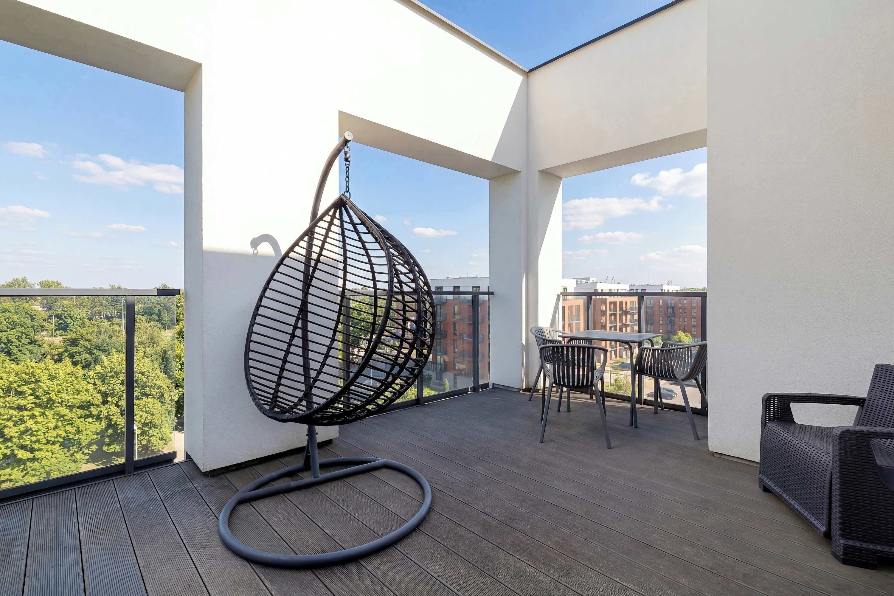 A balcony with a hanging chair, a table with chairs, and a view of greenery and buildings.