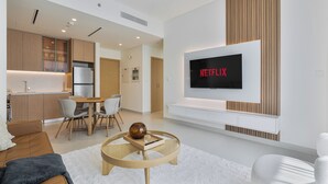 Een moderne woonkamer met een flatscreen televisie aan de wand, een eethoek met een houten tafel en stoelen, en een keuken met houten kasten en een koelkast.