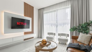 Een moderne woonkamer met een flatscreen televisie aan de muur, een glazen salontafel met een decoratieve schaal, een leren bank met kussens en een groot raam met dunne gordijnen.