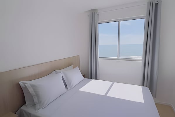 A hotel room with a bed, a view of the sea, and a window with curtains.