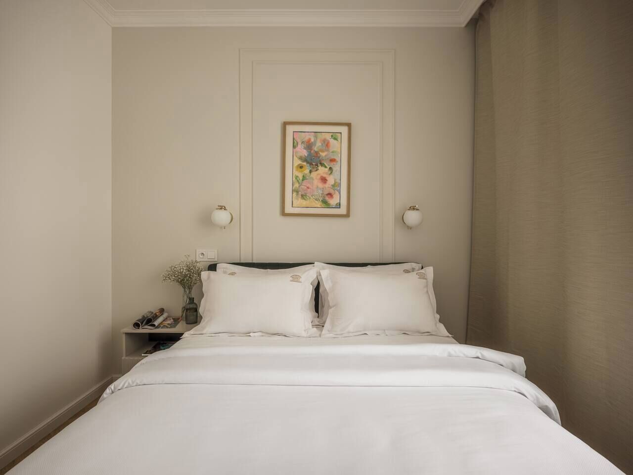 Ein ordentlich bezogenes Bett mit weißen Leinen, ein Nachttisch mit Lampe und ein Blumenbild an der Wand.