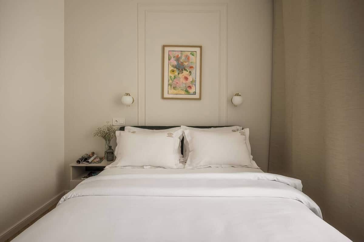 Een net opgemaakt bed met witte lakens, een nachtkastje met een lamp, en een bloemendecoratie aan de muur.