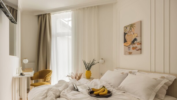 Un lit bien fait, avec une literie blanche, une table de chevet avec une lampe, une chaise et une œuvre d’art accrochée au mur.