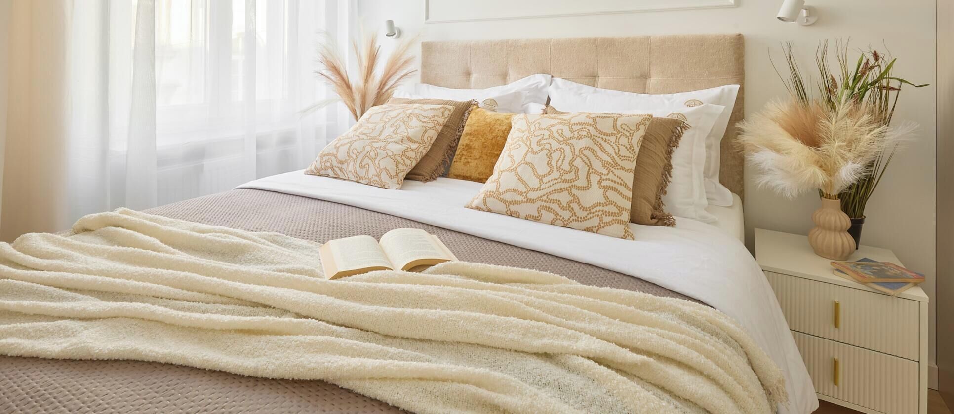 Una cama bien hecha con cabecera color beige, edredón blanco y almohadones estampados. Un libro abierto está sobre la cama, y una mesita de noche blanca con un florero de plantas secas está al lado de la cama.