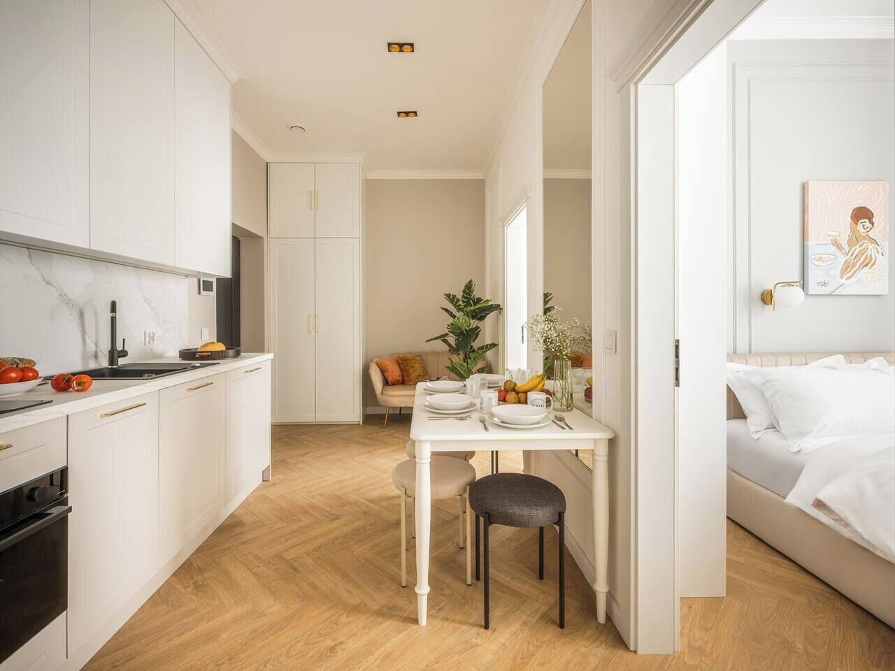 A modern kitchen with white cabinetry, a dining area with a table and chairs, and a bedroom with a bed and a painting.
