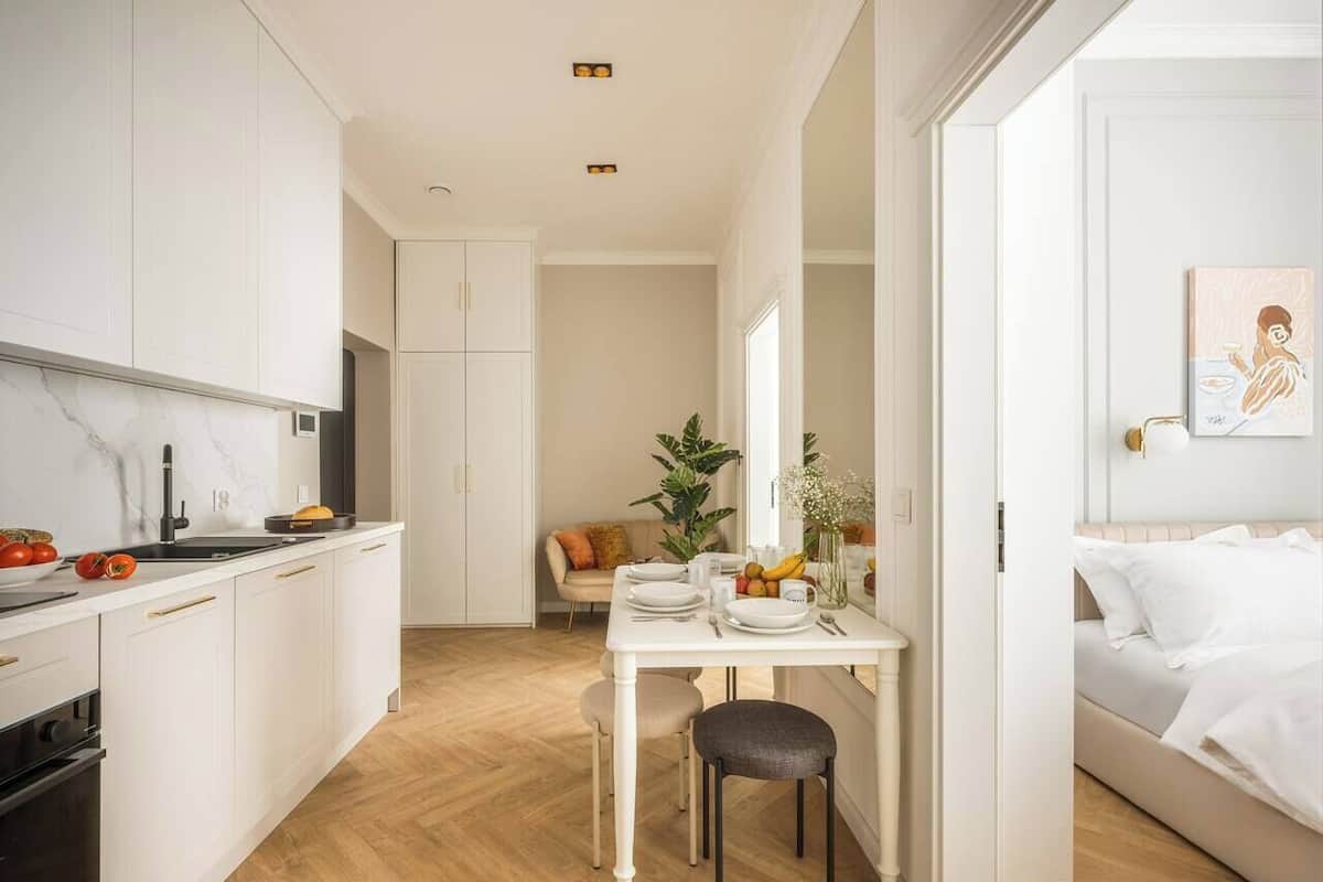 A modern kitchen with white cabinetry, a dining area with a table and chairs, and a bedroom with a bed and a painting.