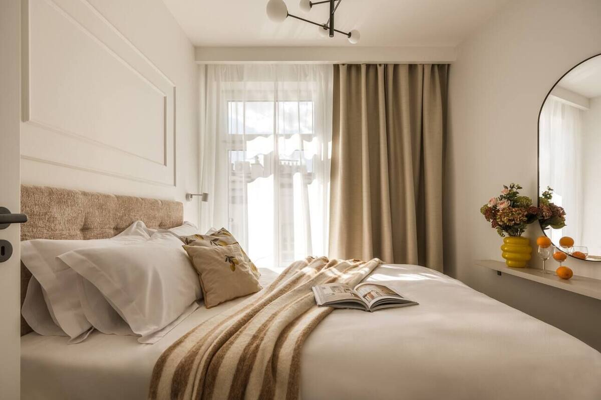 Een keurig opgemaakt bed met een tijdschrift, een vaas met bloemen en een raam met gordijnen.