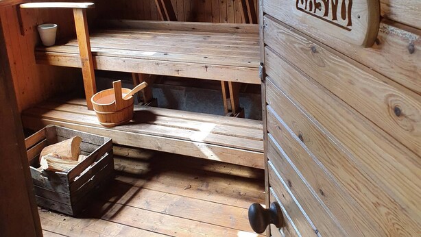 Een houten interieur met een naambordje waarop ‘Bastyu’ staat, houten planken, een houten kruk, een houten emmer en een houten kist.