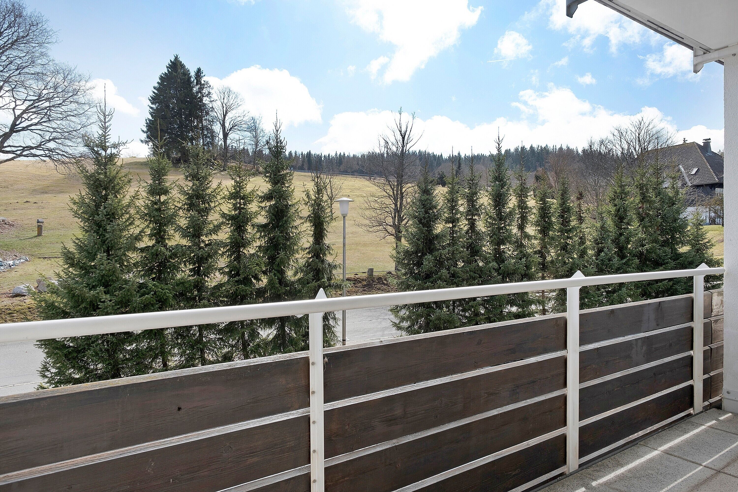 A balcony with a view of a grassy field and trees, a wooden fence, and a house in the background.