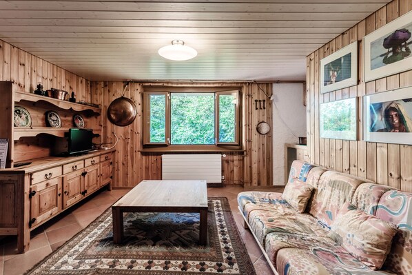 A wooden interior with a sofa, a coffee table, a television, and a window with a view of greenery.