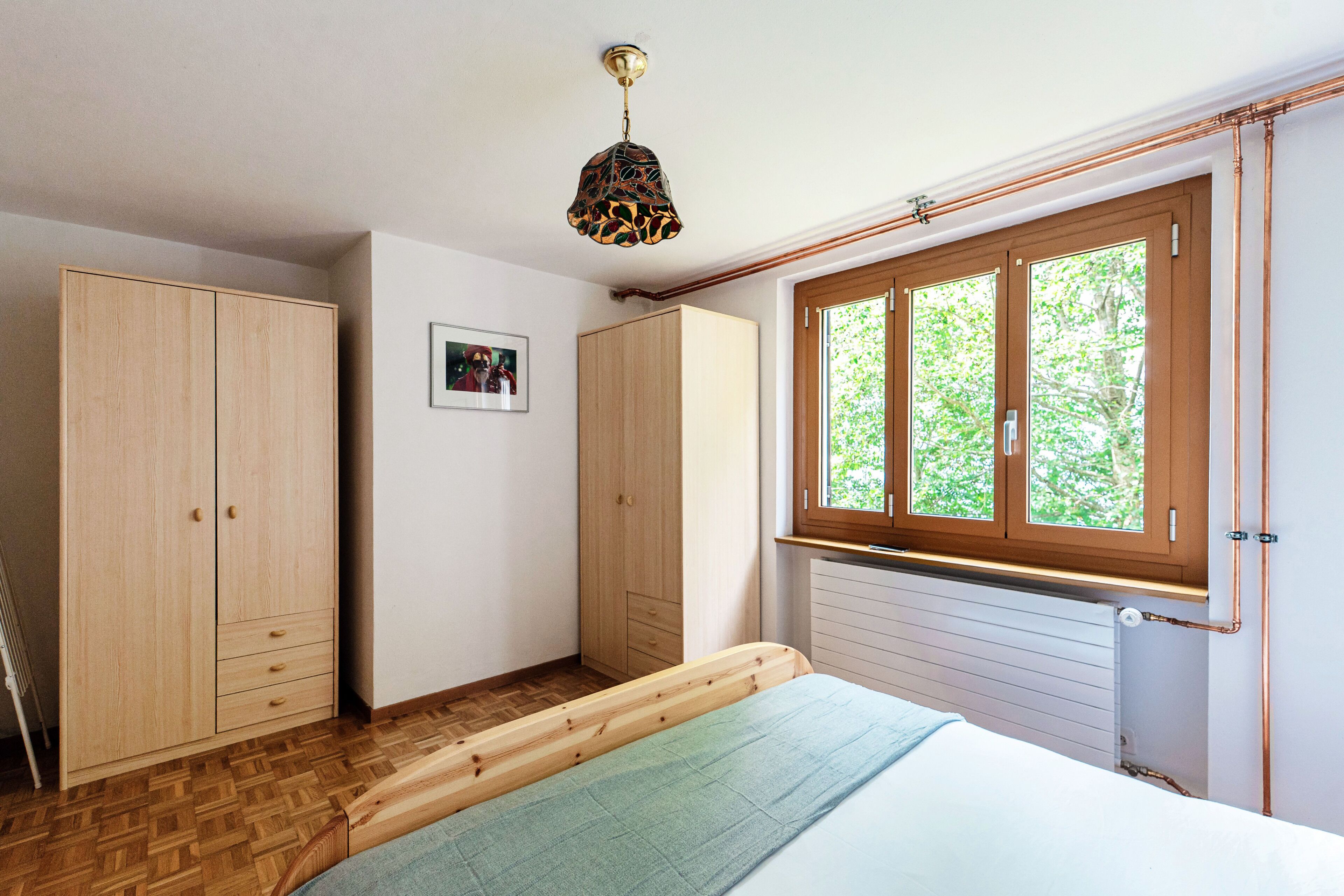 Une chambre avec une armoire en bois, un lit recouvert d’une couverture verte, une fenêtre donnant sur des arbres et un tableau accroché au mur.