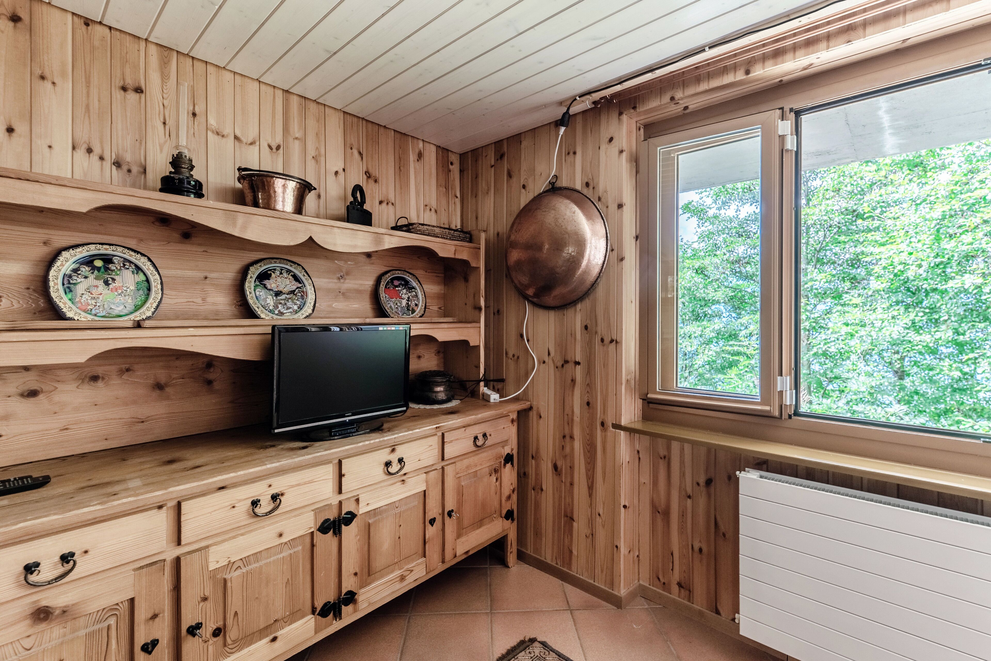 A wooden interior with a television on a cabinet, decorative plates, and a large window with a view of greenery.