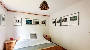Un dormitorio con cama de madera, ropa de cama blanca, una mesita de noche con un objeto rojo y cuadros en la pared.