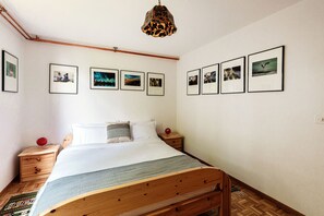 A bedroom with a wooden bed, white bedding, a nightstand with a red object, and framed pictures on the wall.