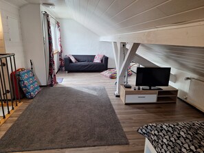 A cozy loft bedroom with a wooden ceiling, a black sofa, a TV on a wooden stand, and a colorful patterned rug.