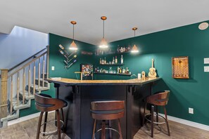 Un coin bar avec un comptoir sombre, trois tabourets et une variété de bouteilles et de verres disposés sur des étagères.