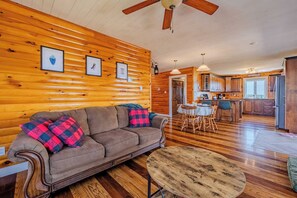 A cozy cabin interior with a wooden log wall, a sofa with plaid cushions, a dining table with chairs, and a kitchen area.