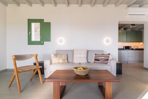 A modern living room with a wooden chair, a sofa with patterned cushions, a wooden table with a fruit bowl, and a kitchen in the background. - Villa Altheon Mykonos (Mykonos)