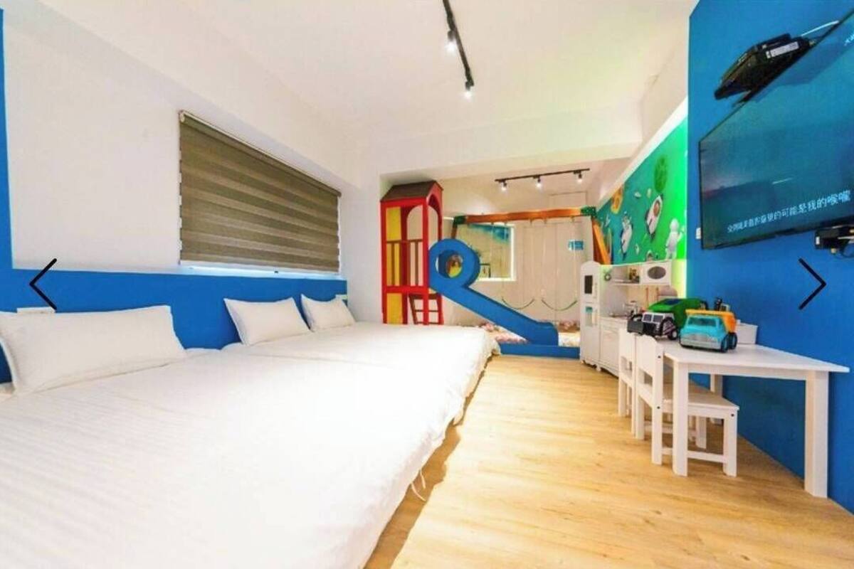 Un dormitorio con cama, escritorio y silla. Se ve una ventana con persianas, un reloj de pared y un televisor montado en la pared.