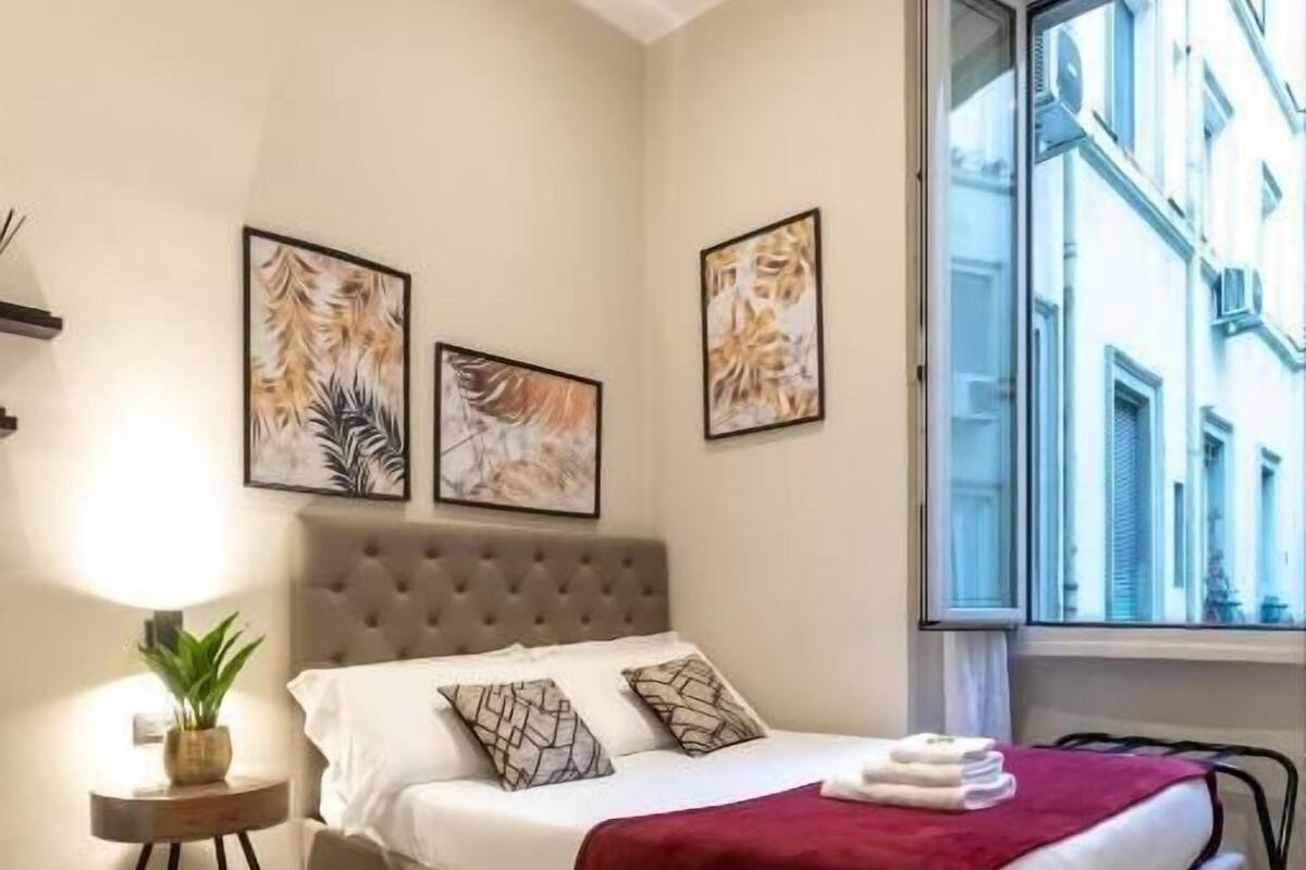 A bedroom with a bed, a bedside table with a plant, and artwork on the wall.