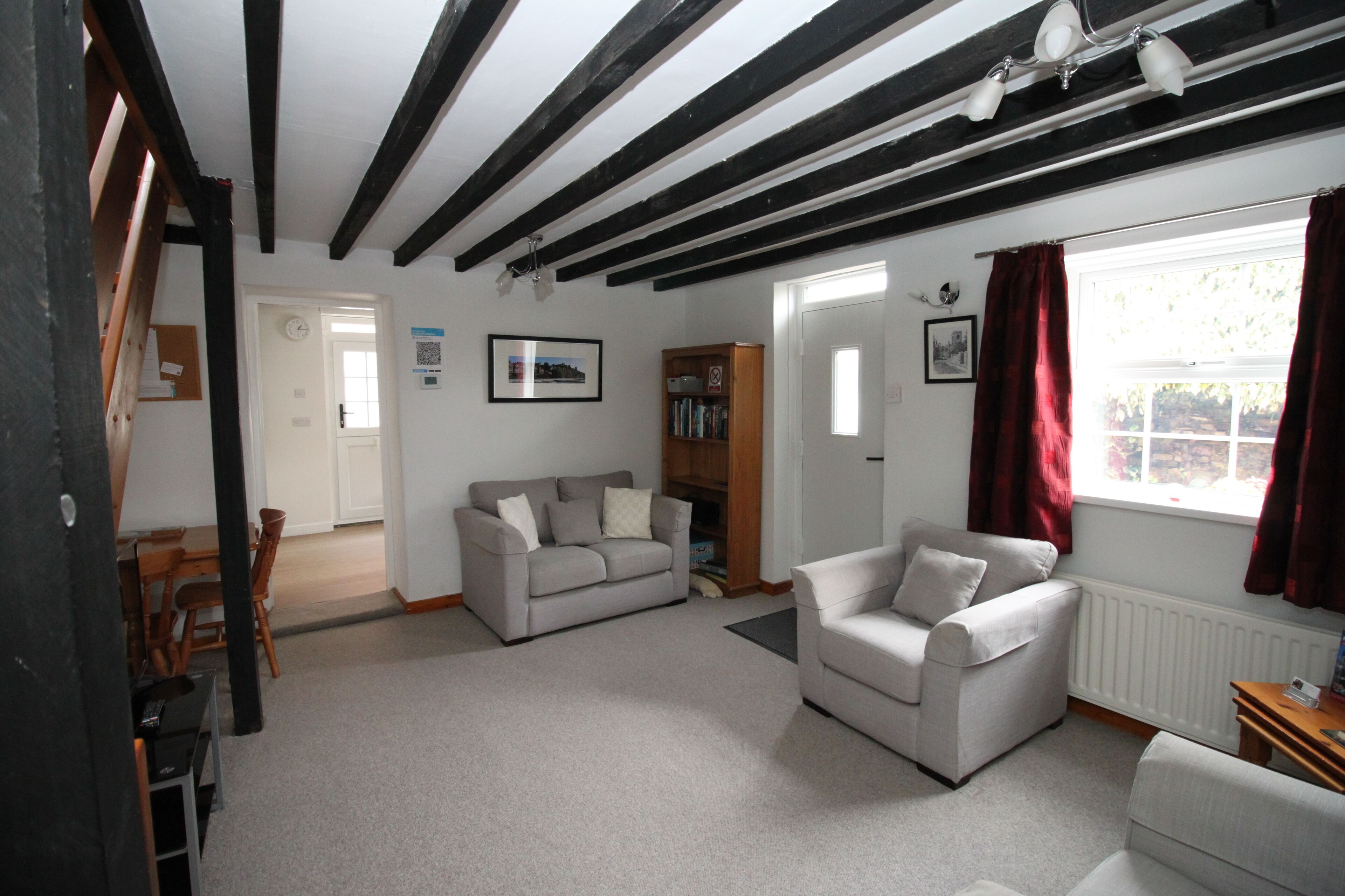 Ein Wohnzimmer mit einer Couch, einem Sessel und einem Holzkabinett. Es gibt ein Fenster mit Vorhängen, eine Tür und eine Decke mit sichtbaren Holzbalken.