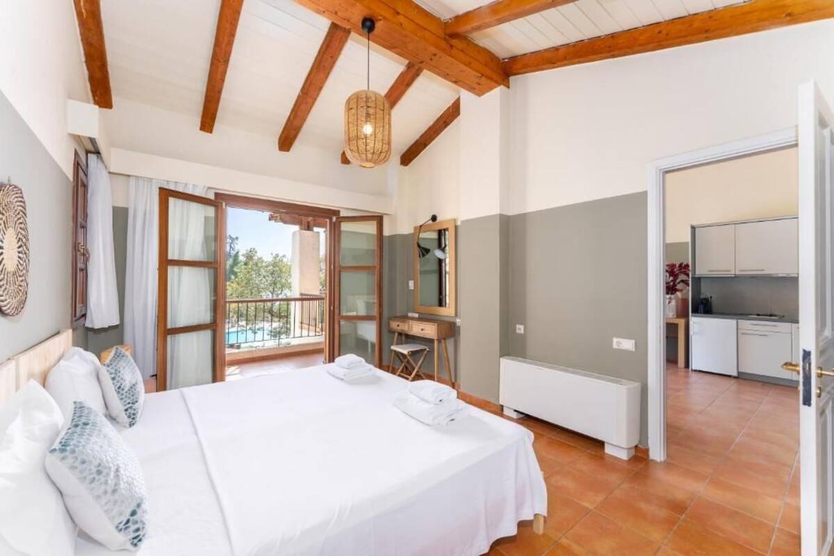 A bedroom with a large bed, a wooden ceiling, a balcony, and a kitchen area.