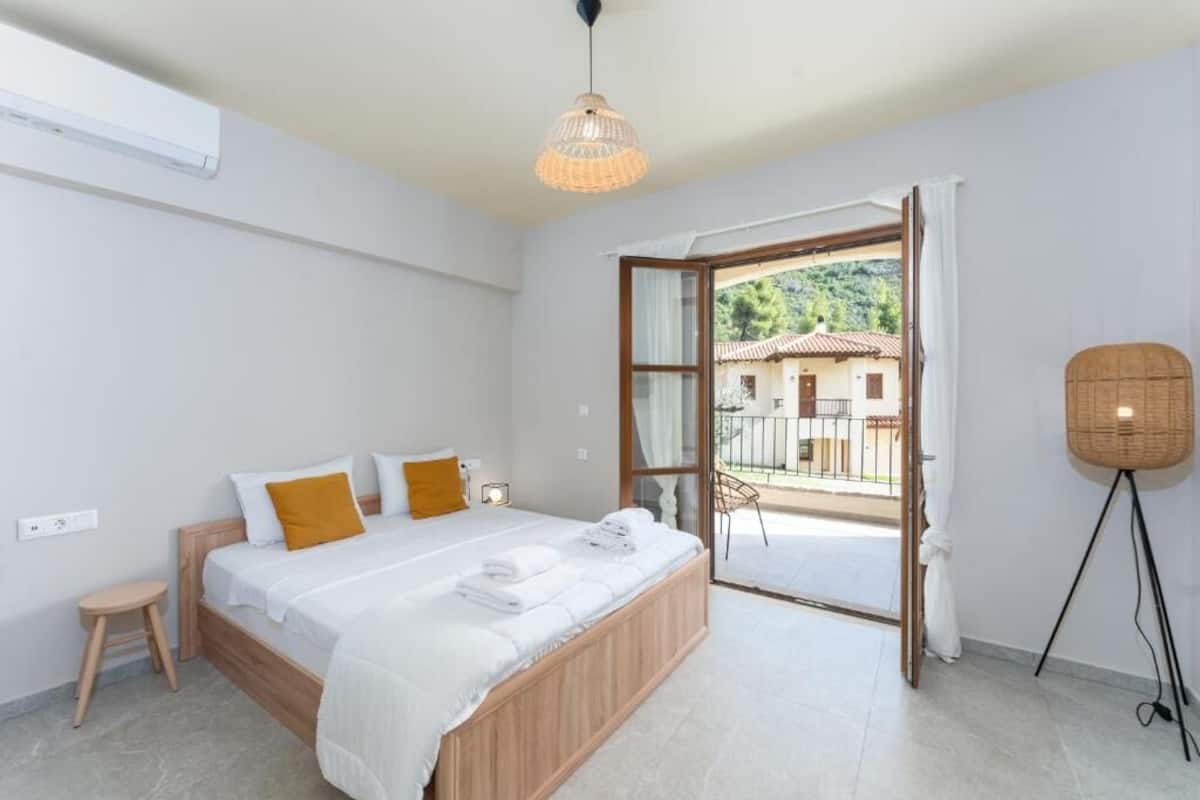A bedroom with a bed, a small stool, a standing lamp, and a balcony with a view of buildings.