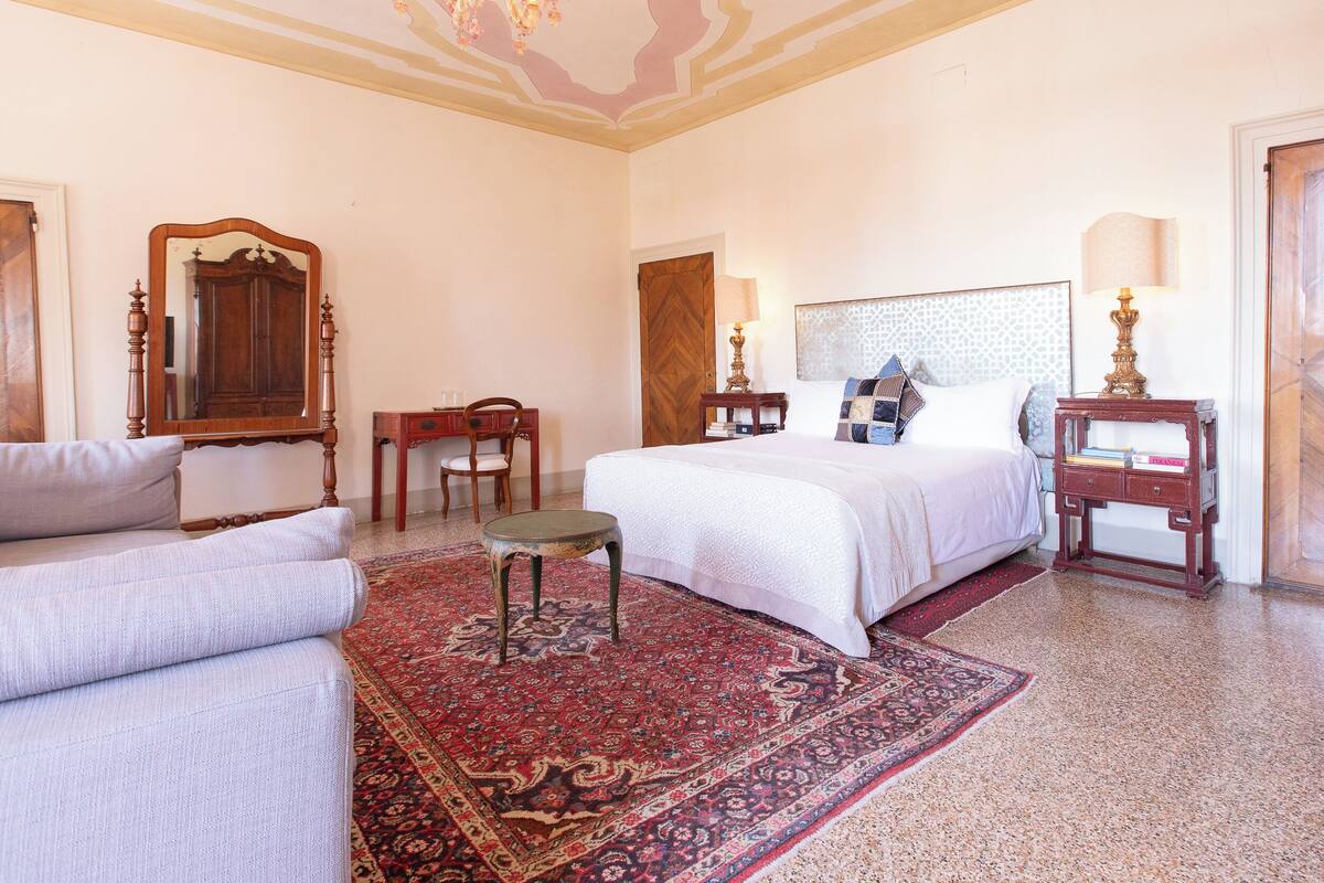 A hotel room with a large bed, a sitting area with a sofa, a small table, a chair, and a decorative rug.