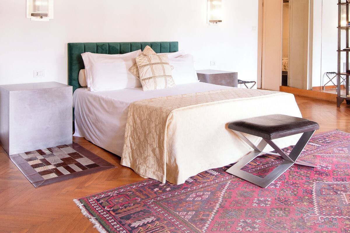 A bedroom with a wooden ceiling, a bed with a green headboard, bedside tables, a bench, and a patterned rug.