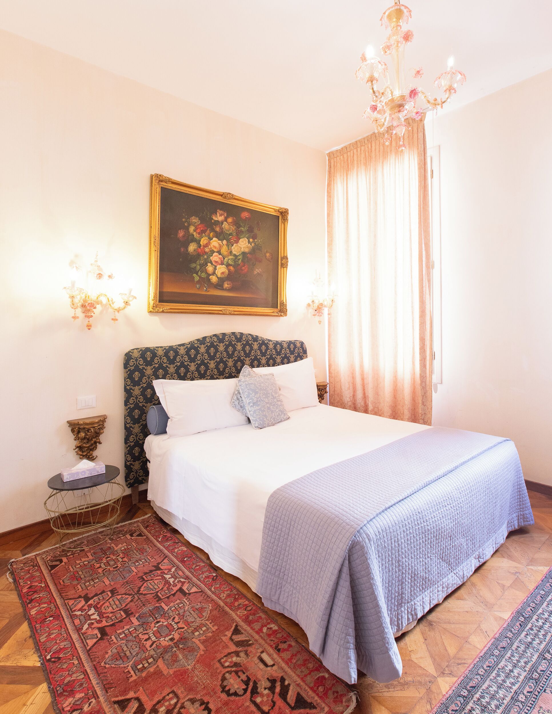 Un dormitorio con una cama, una araña, un cuadro y una alfombra estampada.