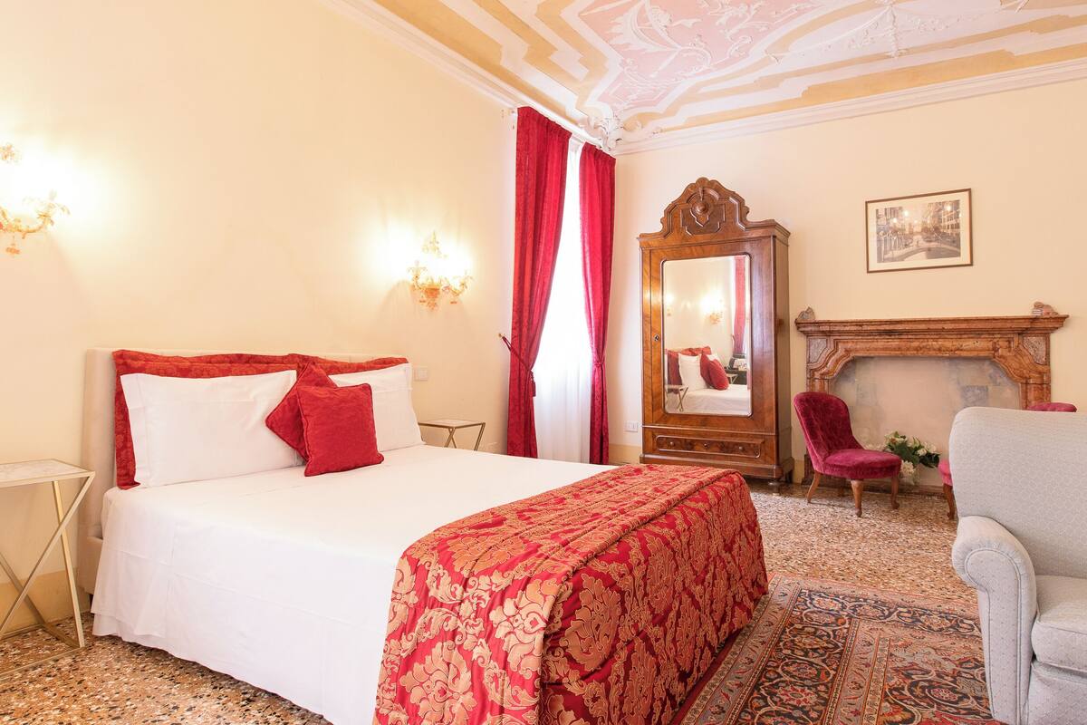 A hotel room with a bed, a red and white patterned bedspread, a wooden wardrobe, a red armchair, a grey armchair, a fireplace, and a chandelier.