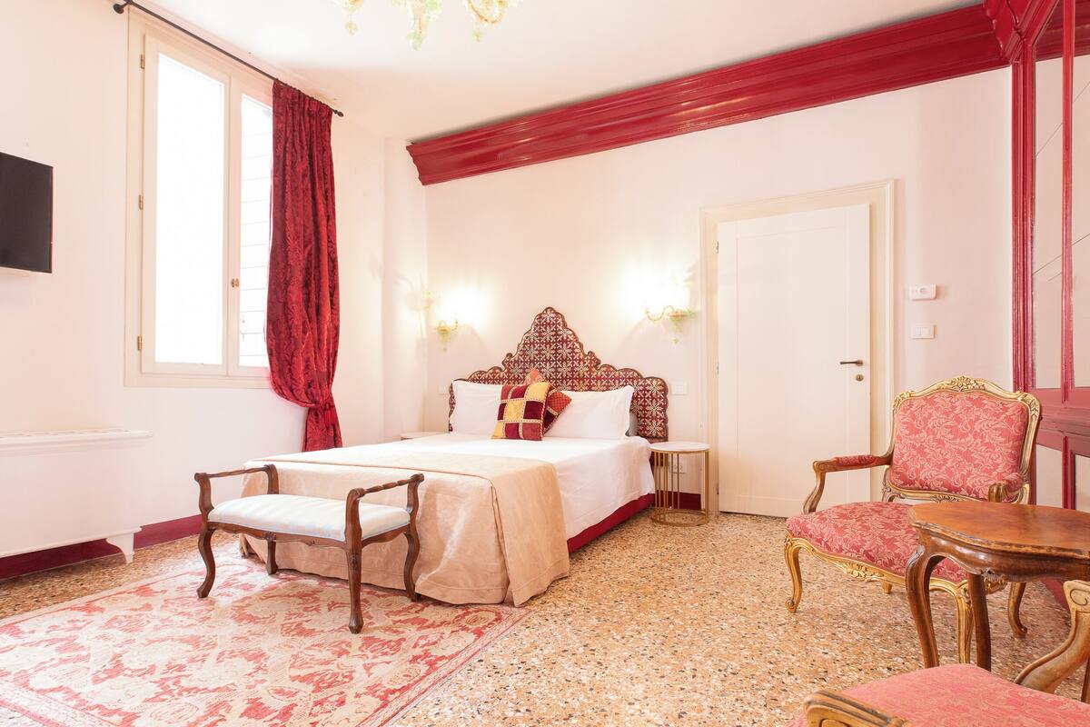 A hotel room with a large bed, a sitting area with a chair and table, a red curtain, and a chandelier.